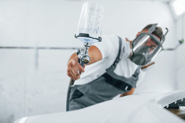Uses painting gun. Caucasian automobile repairman in uniform works in garage