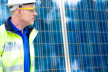 Fototapeta premium Outdoor shot of Caucasian engineer inspect electrical solar panel wearing hardhat , protective eyeglass and safety equipment with smile on his face. Alternative energy and industrial job concept.