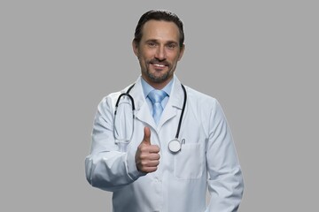 Fototapeta premium Portrait of smiling doctor showing thumb up gesture. Handsome male practitioner with stethoscope expressing success against gray background.