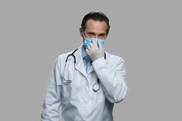 Man doctor in gloves adjusting medical mask. Mature male caucasian doctor wearing protective mask and gloves standing on gray background. Quarantine at hospital, coronavirus concept.