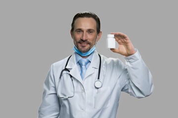 Professional male doctor showing bottle of pills. Attractive mature doctor with surgical mask and stethoscope against gray background.