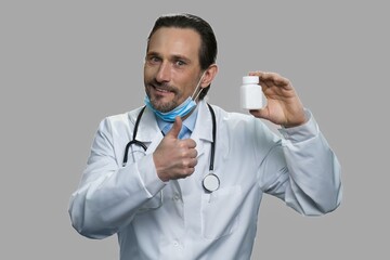 Male doctor showing bottle of pills and thumb up. Cheerful doctor presenting medication against gray background.
