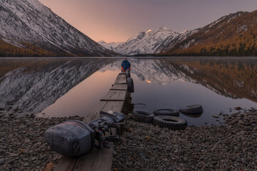 Dreamer. Lonely Man Greets The Dawn Sitting On A Boat Dock On The Shore Of Mountain Lake. Sunrise In Mountains. Old Boat Motor On A Wooden Pier.Reflection Of Snowy Peaks In Calm Water.Siberia, Russia - 403252531