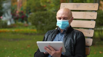 Bald senior man in medical protective mask sits on the bench and holds in hands tablet computer - Powered by Adobe