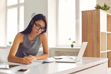 Student in glasses taking notes while watching webinar or listening to online lecture on laptop