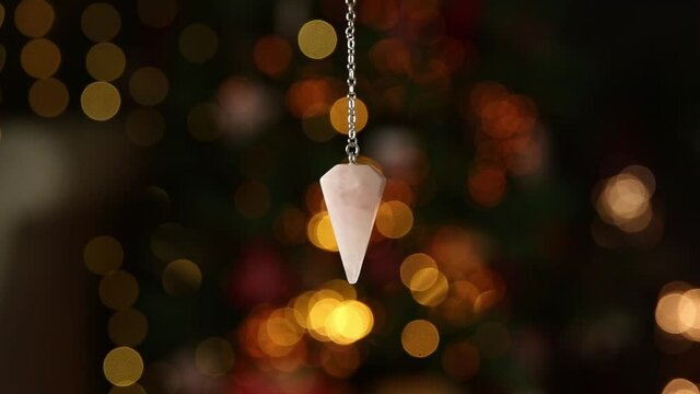 Pink color love rose quartz natural crystal pendulum hanging on silver chain indoors with round yellow bokeh lights on background. New years predictions concept. 
