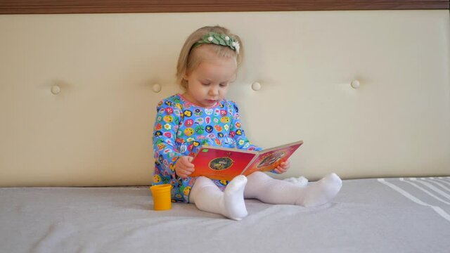 A little girl reads and looks at a picture book. Funny toddler flips through the pages of a book on the bed.