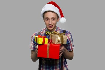 Christmas boy with gift boxes on gray background. Boy in Santa Claus hat holding Christmas gifts and looking at camera.