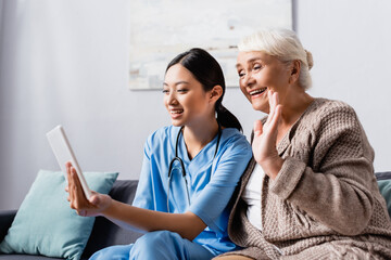 smiling asian nurse holding digital tablet near happy senior woman waving hand during video talk