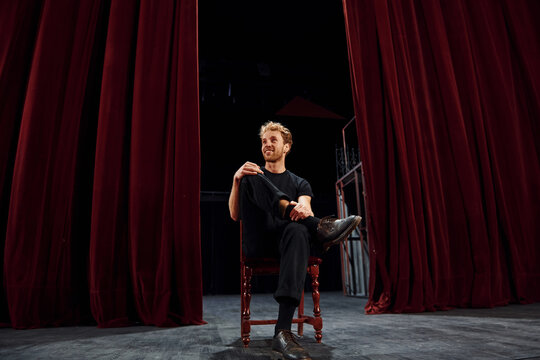 Positive young man sitting on the chair between the curtains on the stage of theater