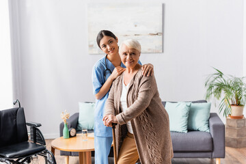 smiling asian nurse and elderly woman with walking stick looking at camera in nursing home