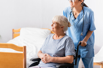Obraz premium young nurse near smiling aged woman sitting in wheelchair and looking away