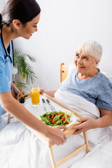 Obraz na płótnie Canvas smiling asian nurse holding tray with breakfast near happy elderly woman in bed