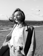 Portrait of a laughing young woman in coat, white shirt and jeans enjoying of bright sunlight standing on beach in front of sea. Girl having fun outside. Urban style and fashion. Casual outfit