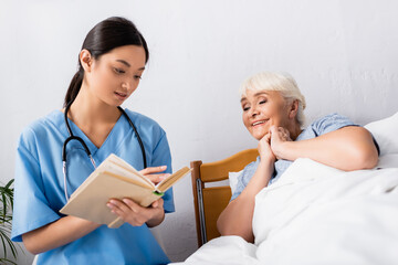 Fototapeta premium asian nurse reading book to cheerful elderly woman in hospital