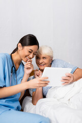 Obraz na płótnie Canvas excited asian nurse and senior woman laughing while using digital tablet together