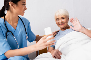 Fototapeta premium young asian nurse holding mobile phone near cheerful aged woman waving hand during video call