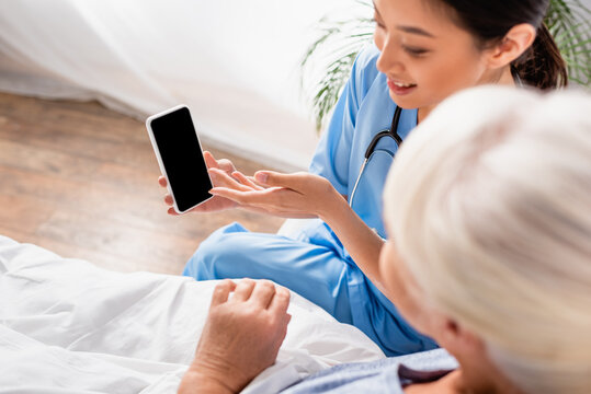 Overhead View Of Smiling Asian Nurse Pointing At Mobile Phone Near Senior Woman On Blurred Foreground