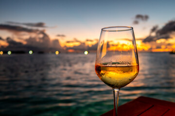 Glass of white wine on a table with dramatic sunset in the background and lights on horizon, Maldives.