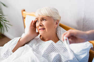 Obraz premium nurse holding thermometer near sick senior woman suffering from headache, blurred foreground