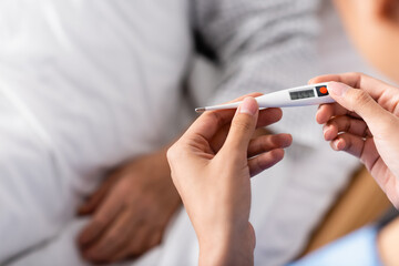 partial view of nurse holding thermometer near aged patient, blurred background
