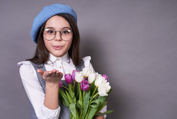 Beautiful fashionable Caucasian lady in a stylish blue hat in a beret and glasses holds flowers tulips in his hands on a gray background.