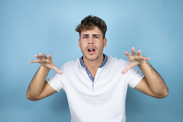 Young handsome man wearing a white t-shirt over blue background smiling funny doing claw gesture as...