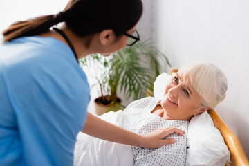 Fototapeta premium young nurse touching cheerful aged woman lying in hospital, blurred foreground