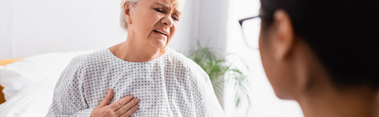 senior woman touching chest while suffering from heart pain near nurse on blurred foreground, banner