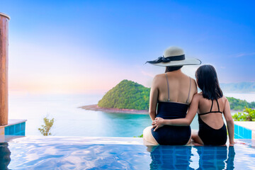 Asian women in black swimsuit with her daughter siting on side of infinity pool relaxing at sunset time  with sea view during vacation retreat. Family happy and enjoy with holiday summer travel time.