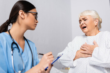 Fototapeta premium aged woman touching chest while suffering from asthma attack near asian nurse writing on clipboard, blurred foreground
