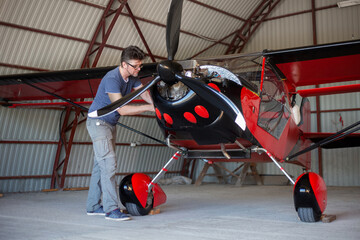 Repairman looking over engine in small aircraft from below. Small red airplane in hangar. Repaiper...
