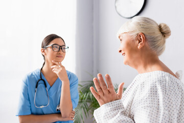 young asian nurse in eyeglasses listening to aged woman talking and gesturing in hospital, blurred background