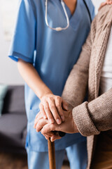 cropped view of nurse touching hands of aged woman standing with walking stick, blurred background