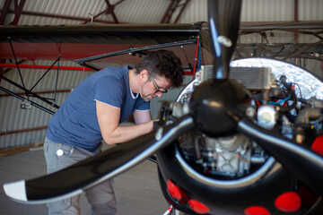 Repairman looking over engine in small aircraft from below. Small red airplane in hangar. Repaiper holding instruments in the hand near the plane engine. Small aviation.