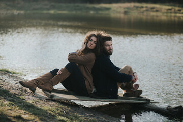 two caucasian lovers sitting on the pier by the lake. Young couple is hugging on autumn day outdoors. A bearded man and curly woman in love. Valentine's Day. Concept of love and family.