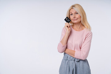 Dreamy aged woman with credit card in casual outfit isolated over grey background