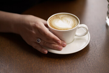 Female hands holding a white cup of delicious coffee with foam over wooden table, side view. Valentine's Day coffee