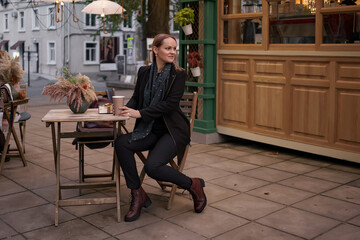 Beautiful Caucasian female is drinking delicious coffee and looking aside while sitting in the street cafe along and waiting for dessert
