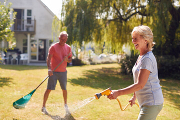 Retired Couple At Work Watering Plants With Hose And Tidying Garden With Rake At Home