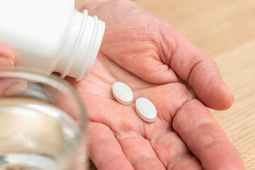 White pills of painkiller or antibiotic for treatment on senior woman hand palm, glass with water, medicines and vitamin supplements concept,close-up view