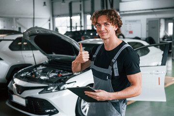 With notepad in hands. Adult man in grey colored uniform works in the automobile salon
