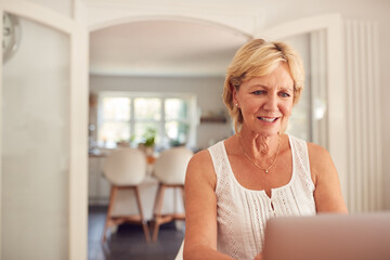 Retired Woman At Home In Kitchen Shopping Online And Managing Domestic Finances Using Laptop