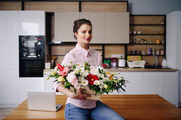 Portrait beautiful woman holding a flower composition posing on white kitchen at home with laptop. Florist presenting a basket with colorful flowers