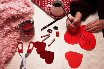 Preparing for the holiday Valentine's Day. The girl makes heart-shaped self-congratulatory cards. Woman signs a postcard with a brush. Cardboard hearts, pink cup, pastel and watercolors