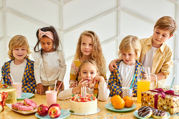 multi-ethnic friendly kids celebrating birthday of friend girl at home having tasty cake, party time, in decorated room.