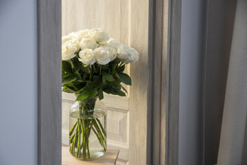 A bouquet of yellow white roses are in a round glass vase made of corrugated glass on a high wooden stool in the doorway as an interior decoration.