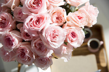 Bouquet of pink roses in a glass vase with a white teapot and two cappuccino cups on a tray on a coffee table among books