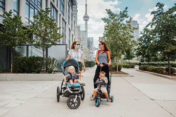 Two Caucasian moms with strollers and kids walking together in Toronto city Canada. Women in face masks with children outdoor. Friends talking on street keeping social distance. A new normal.