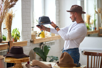 male hats maker standing at his atelier workshop, holding perfectly made hat for customers....